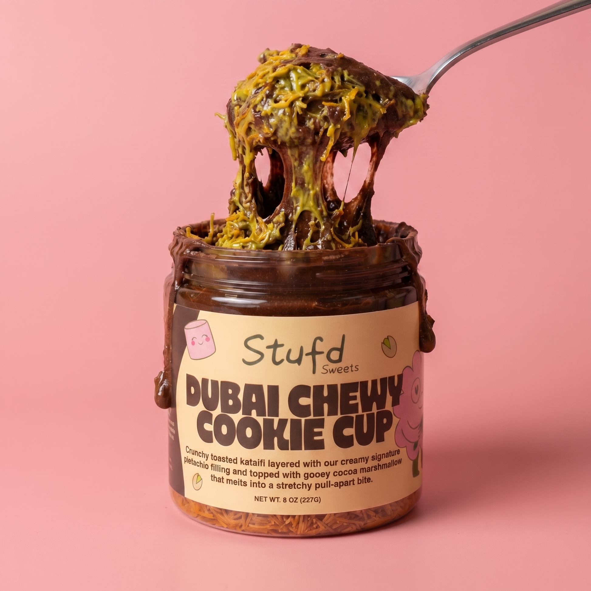 Jar of 'Dubai Chewy Cookie Cup' with a spoonful of cookie on a pink background