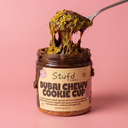 Jar of 'Dubai Chewy Cookie Cup' with a spoonful of cookie on a pink background